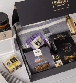 A gift pack featuring a grey blanket, gourmet snacks, nut mix, crackers, and a jar of preserve, all in a black box.