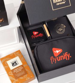 A black gift box contains a branded tech kit, featuring a black pouch, notebook, pen, and snacks.