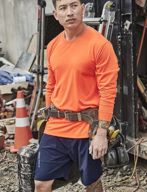 An orange long sleeve safety tee is worn by a man with a tool belt, set against a cluttered worksite background.