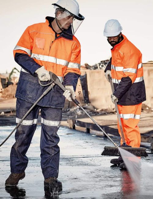 Two workers in high visibility orange and navy jackets, wearing helmets and gloves, operate equipment on a worksite.