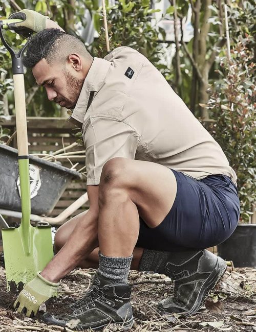 A man in a beige shirt and navy shorts kneels in a garden, working with a green shovel and wearing gloves and boots.