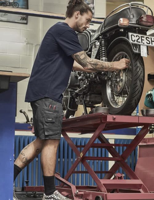 A man in navy blue shorts works on a motorcycle, showcasing rugged cooling stretch cargo shorts with pockets.
