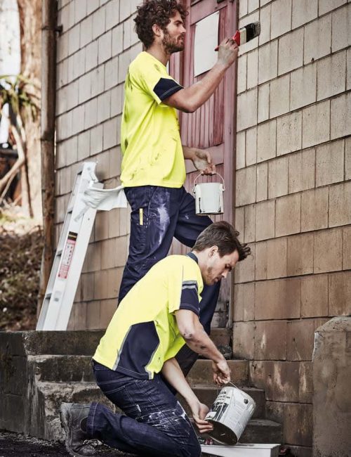 A bright yellow men's high visibility T-shirt worn by two men painting a wall, one standing and one kneeling.