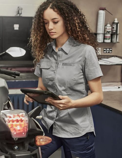 Lightweight grey short sleeve shirt for women, featuring a front button closure and a logo, displayed in a workshop setting.
