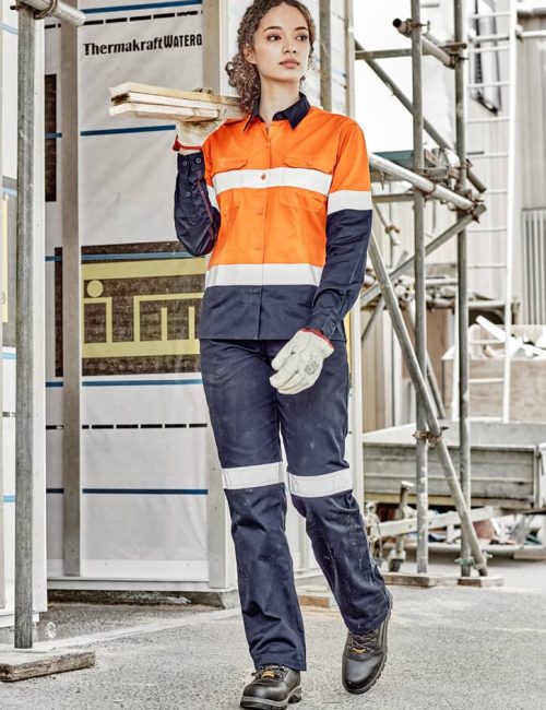 A woman in an orange and navy long sleeve industrial shirt, holding timber, wears work gloves and sturdy boots.
